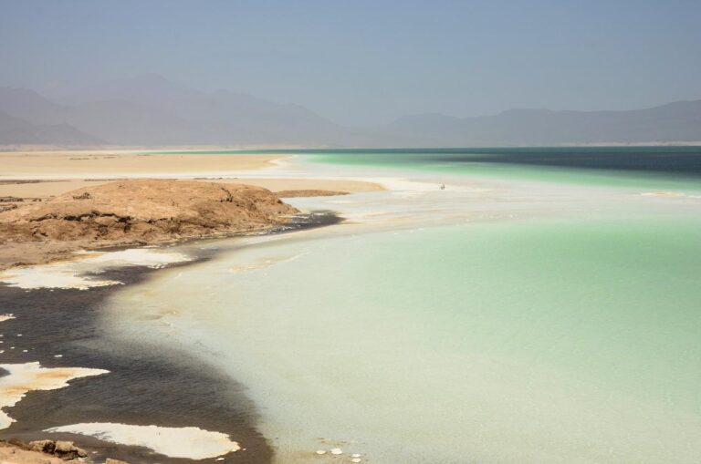 A stunning landscape with a large rock formation, turquoise water, sandy beaches, and distant mountains under a hazy sky.