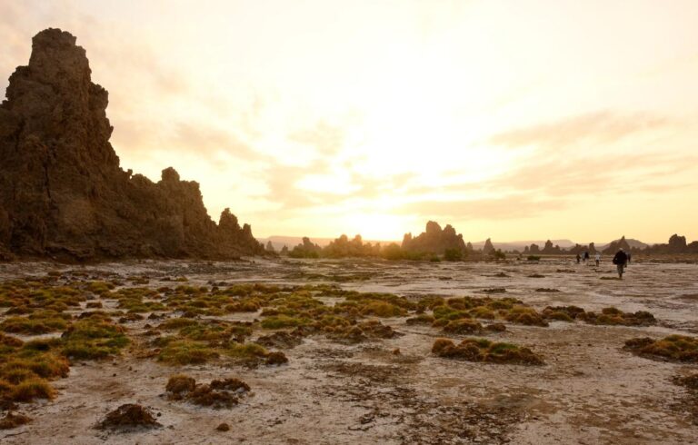 A picturesque landscape of tufa towers and sparse vegetation under a golden sunset sky, with several people walking in the distance.