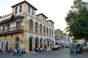 Djibouti City Quarters, Mosque and Market Cultural Day Tour A street scene features aged yellow and white buildings with arched walkways, people sitting and standing, cars, and a tree.