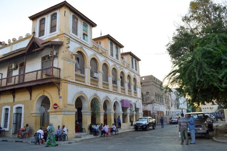 A street scene features aged yellow and white buildings with arched walkways, people sitting and standing, cars, and a tree.