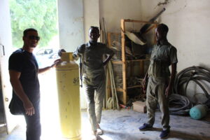 Somalia Project Deployment or Scouting & Survey Service Three men stand indoors near a yellow cylinder, with two men wearing military-style clothing and one wearing a black t-shirt and sunglasses.