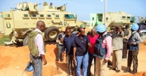 Somalia Private Security Services A group of people, some wearing blue helmets and vests, stand near a military vehicle with "AU" written on it, next to a large pit in a sandy area.