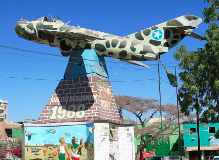 A monument featuring a camouflaged warplane atop a pyramid-shaped base with inscriptions and murals stands against a clear blue sky.