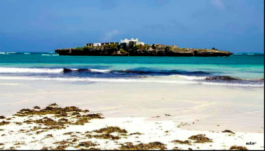 A scenic view of a tropical beach with white sand, turquoise water, and an island with a white building on it under a blue sky.