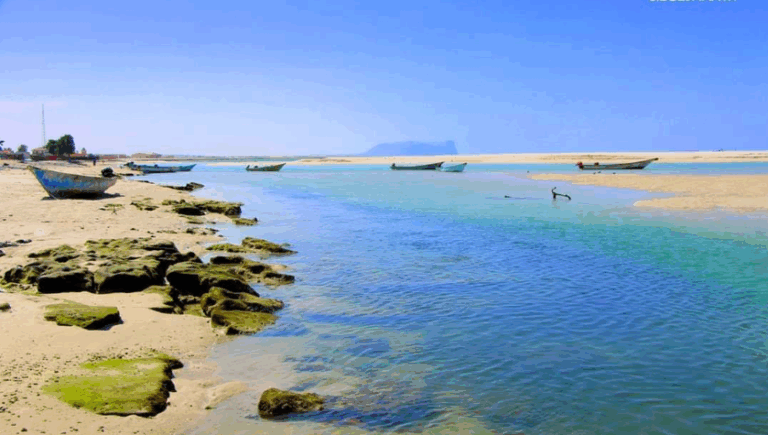 A serene coastal view showcases boats resting in shallow, turquoise waters against a backdrop of sandy beaches and a clear blue sky.