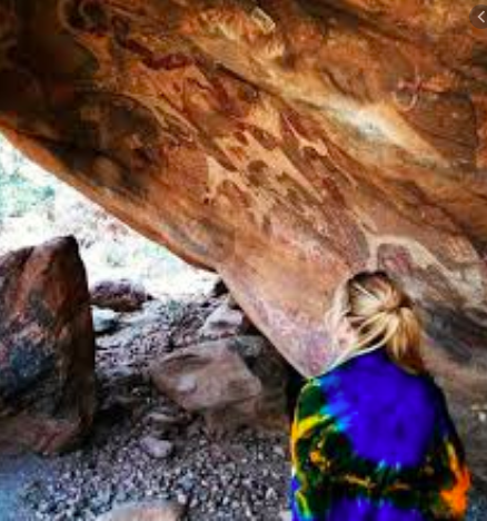 A woman stands in a cave adorned with ancient rock paintings, her colorful shawl contrasting with the natural tones of the stone.